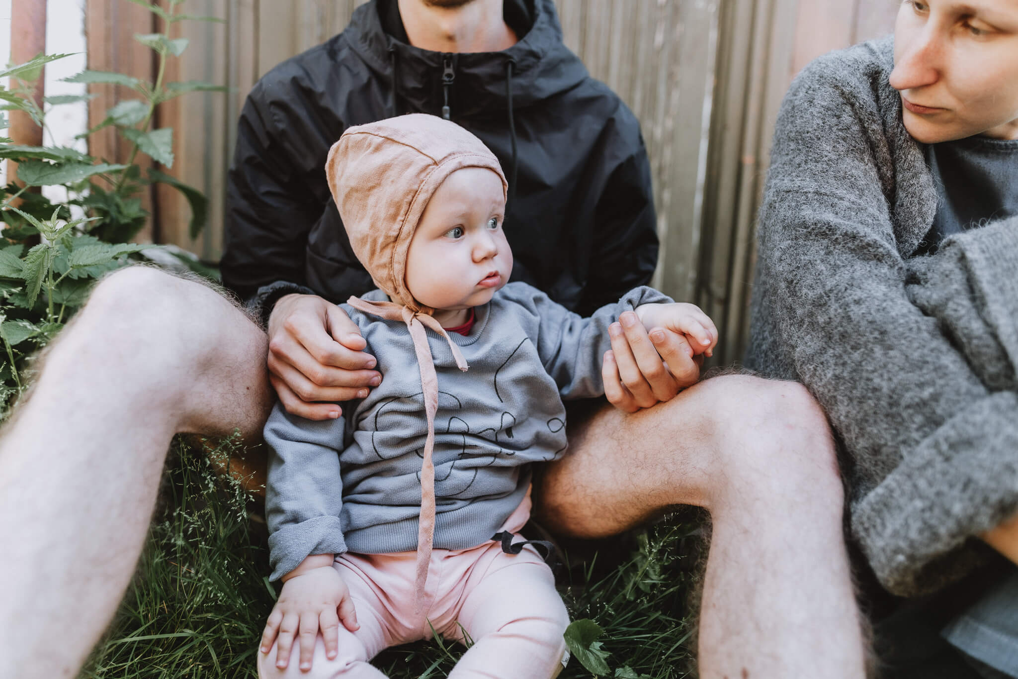 Portreefoto mütsiga beebist, kes istub õues isa süles, ema on kõrval.